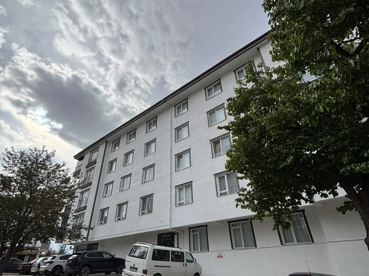 Bulutlu gökyüzü altında binanın uzun beyaz yan cephesi ve önündeki kilit taşı kaplı otopark