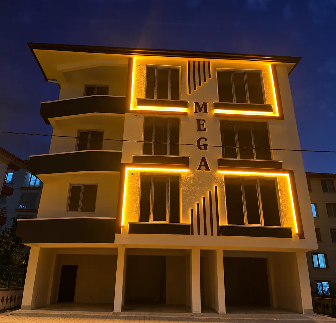 Gece mavi gökyüzü altında pencere çerçeveleri sarı LED ışıklarla aydınlatılmış, cephesinde MEGA harfleri bulunan dört katlı bina